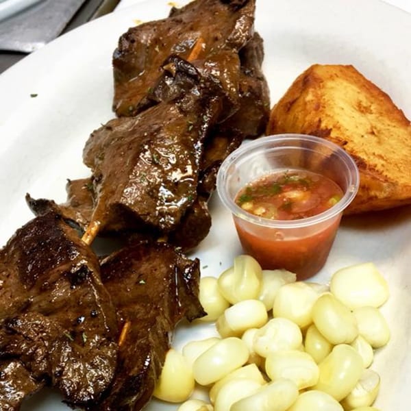 Peruvian anticuchos, a cup of aji sauce, fried potato, and choclo (Peruvian corn) on a white plate.