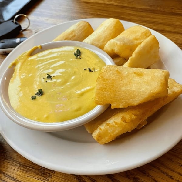 Fried yuca sticks with a side of yellow huancaína sauce, garnished with herbs, on a white plate.